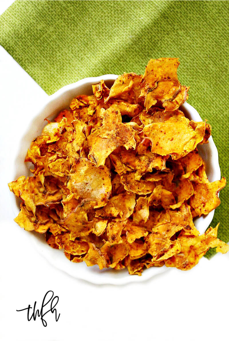 A vertical overhead image of a white bowl filled with Gluten-Free Vegan Dehydrated Sweet Potato Chips on a green napkin on top of a solid white background