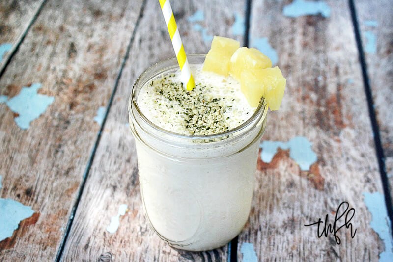 A mason jar filled with Pineapple and Banana Hemp Smoothie garnished with pineapple chunks and straw on a weathered wooden surface