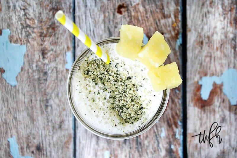 Overhead view of a glass of Pineapple and Banana Hemp Smoothie garnished with a straw and pineapple chunks on top of a weathered wooden surface