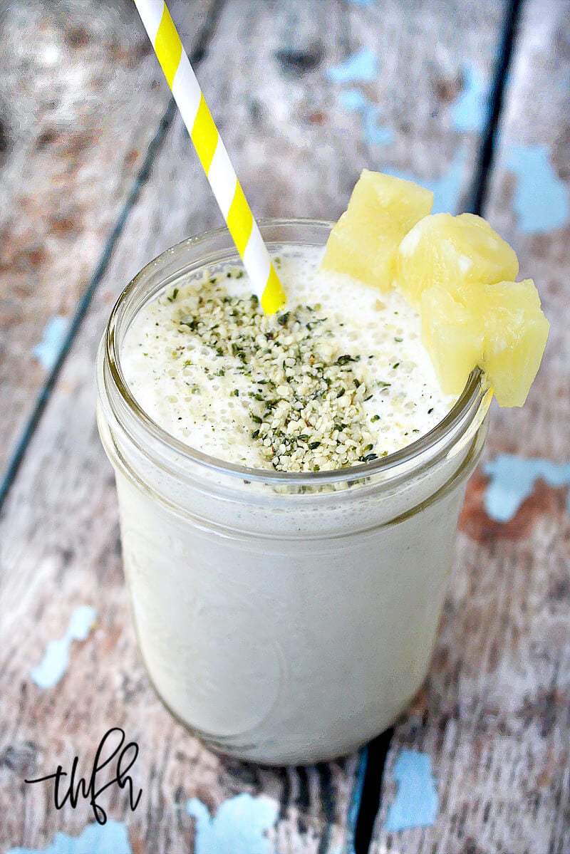 Vertical image of a glass of Pineapple and Banana Hemp Smoothie with a yellow and white straw and pineapple chunks on a weathered wooden surface