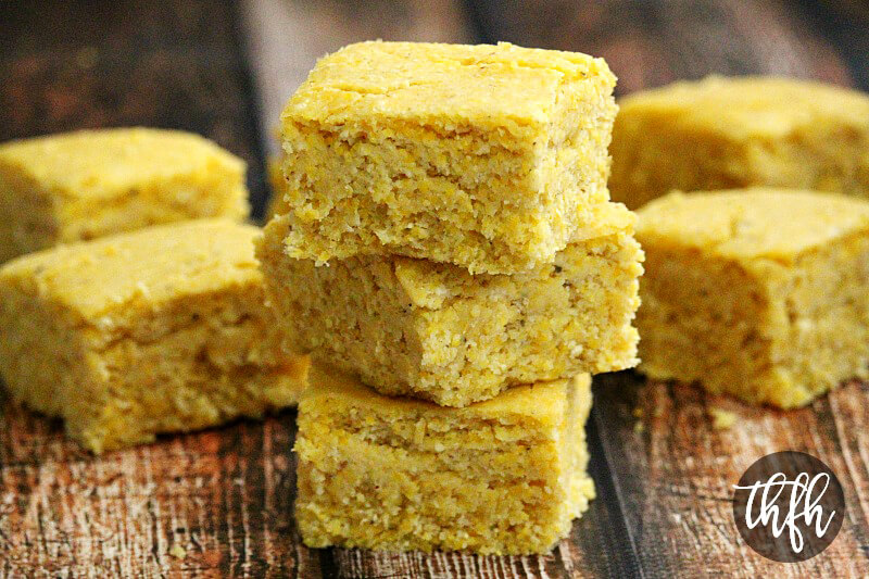 Stack of three sliced squares of The BEST Homemade Vegan Cornbread on a weathered wooden surface with more cornbread slices in the background