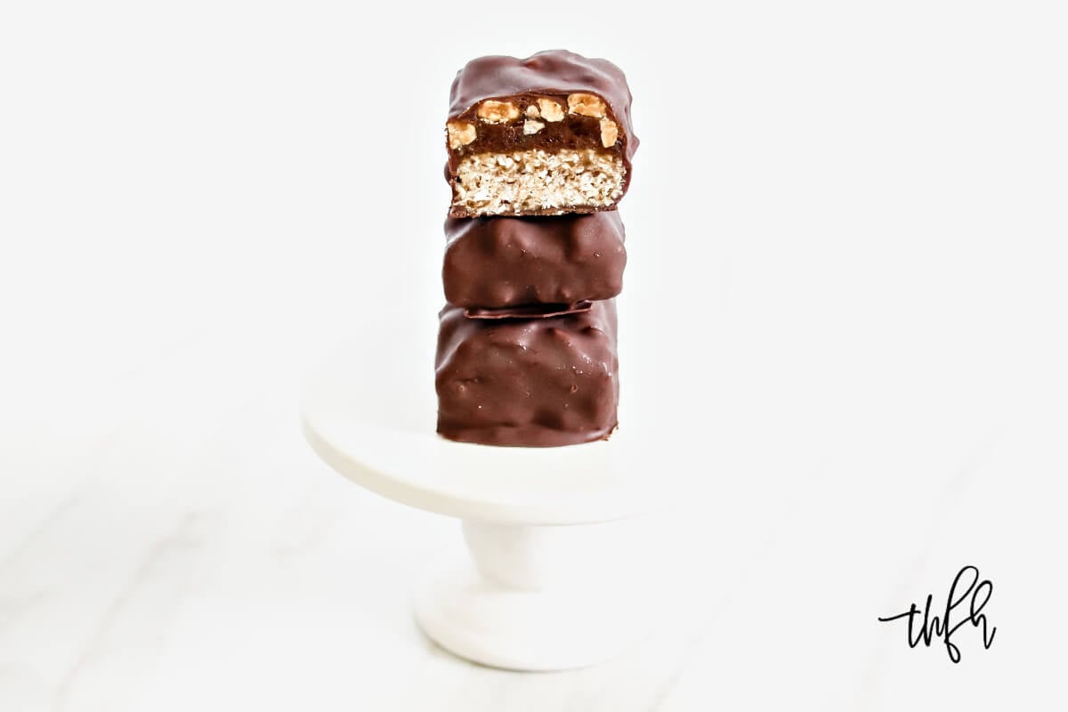 A stack of three homemade snickers bars on a small white pedestal on a solid white background