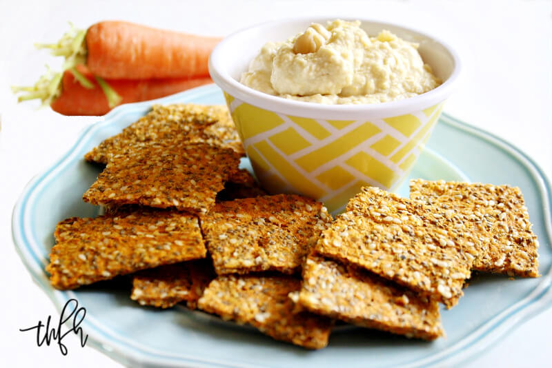 Horizontal image of a blue plate filled with Gluten-Free Vegan Raw Carrot Pulp and Flax Seed Crackers next to a bowl of hummus on a white background