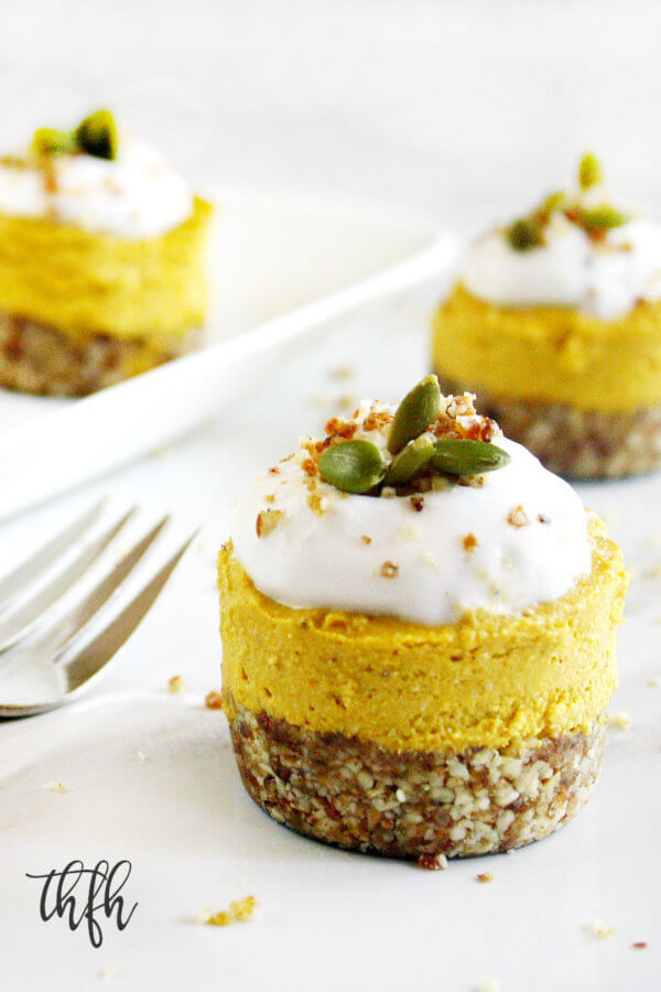 Three individual Gluten-Free Vegan No-Bake Pumpkin Mini Cheesecakes topped with homemade whipped coconut topping on a white marble surface
