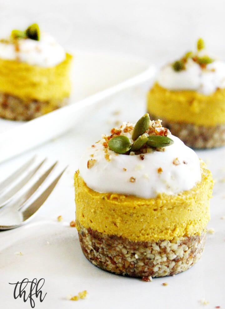 Three individual Gluten-Free Vegan No-Bake Pumpkin Mini Cheesecakes topped with homemade whipped coconut topping on a white marble surface