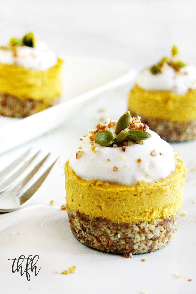 Vertical image of three Gluten-Free Vegan No-Bake Pumpkin Mini Cheesecakes topped with homemade whipped coconut cream with one prominently in the forefront