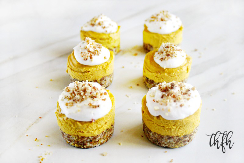 Six Gluten-Free Vegan No-Bake Pumpkin Mini Cheesecakes topped with homemade whipped coconut topping lined up vertically on a white marble surface