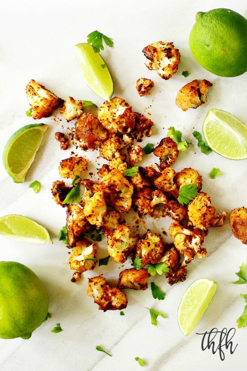 Vertical overhead image of Gluten-Free Vegan Roasted Cauliflower with Chipotle and Lime scattered on a white marble surface with sliced limes