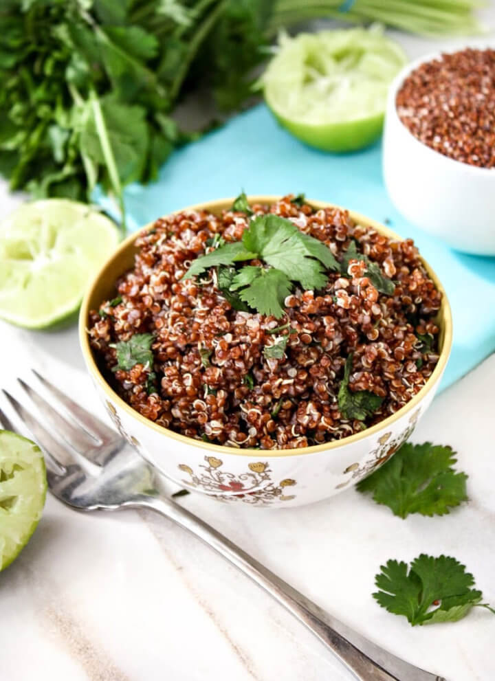 Vertical image of a small decorative bowl filled with Red Quinoa with Cilantro and Lime with a fork next to the bowl and squeezed limes to the side