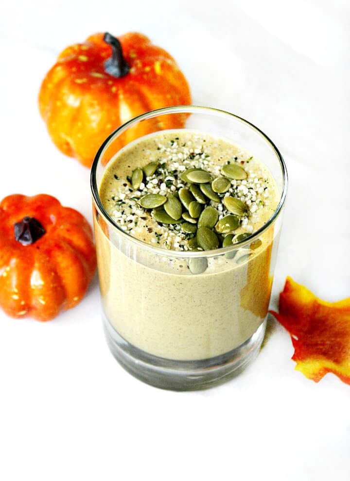 Vertical image of a glass filled with The BEST Vegan Pumpkin Protein Smoothie on a solid white marbled surface with two small pumpkins in the background