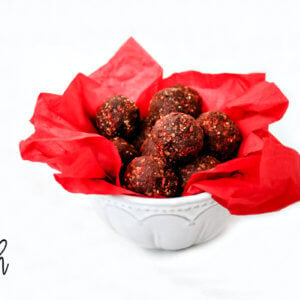 A decorative white bowl lined with red paper filled with chocolate truffles on a solid white background