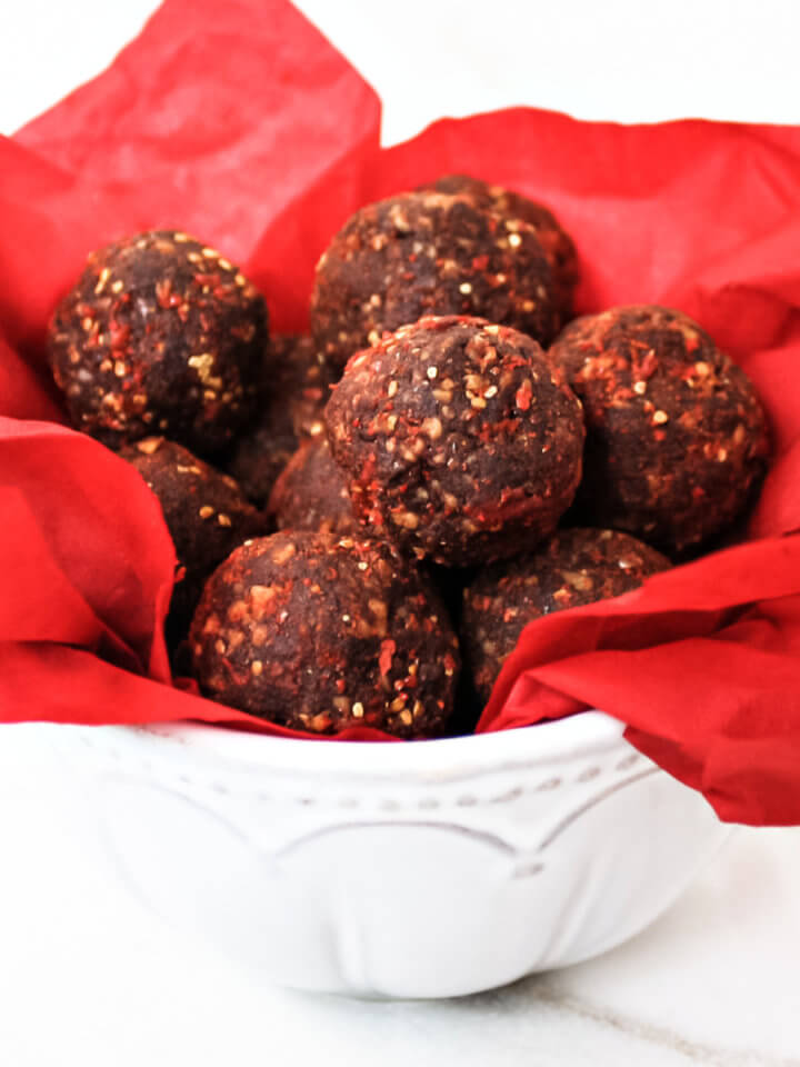 A decorative white bowl lined with red paper filled with chocolate truffles on a solid white background