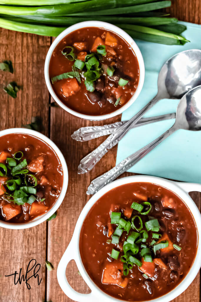 Overhead image of a large ceramic pot filled with chili next to two smaller white bowls filled with chili on a weathered wooden surface
