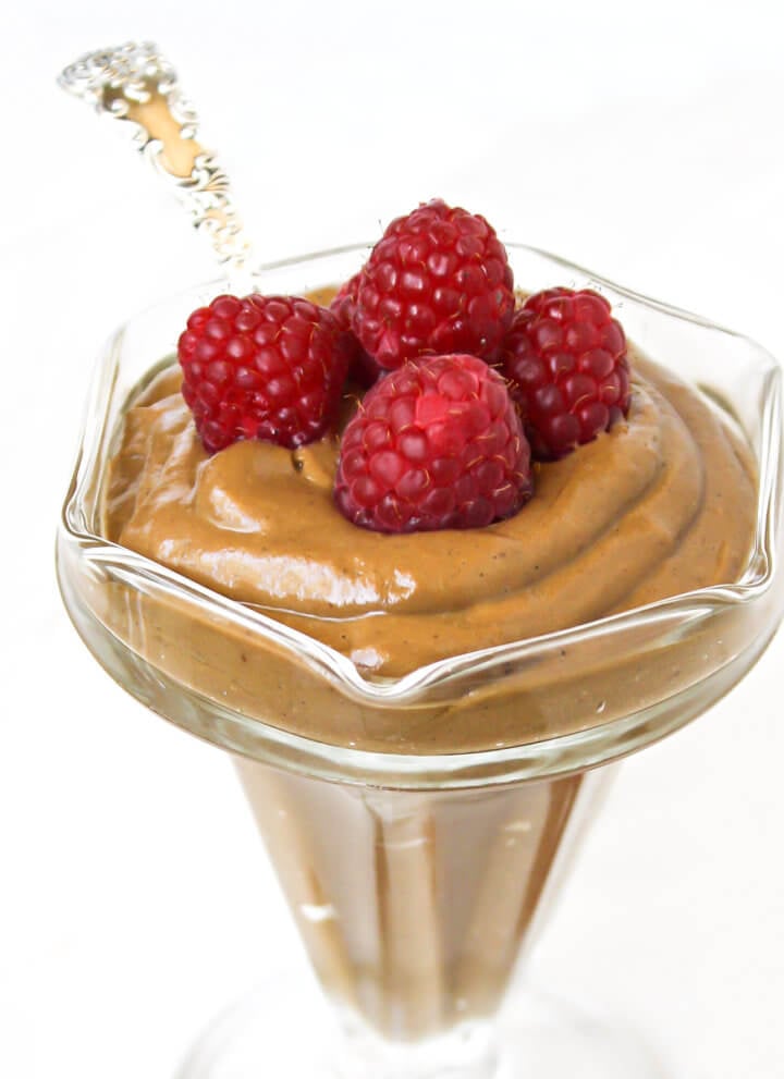 Square image of a glass dish filled with chocolate pudding topped with multiple raspberries with a silver spoon inserted