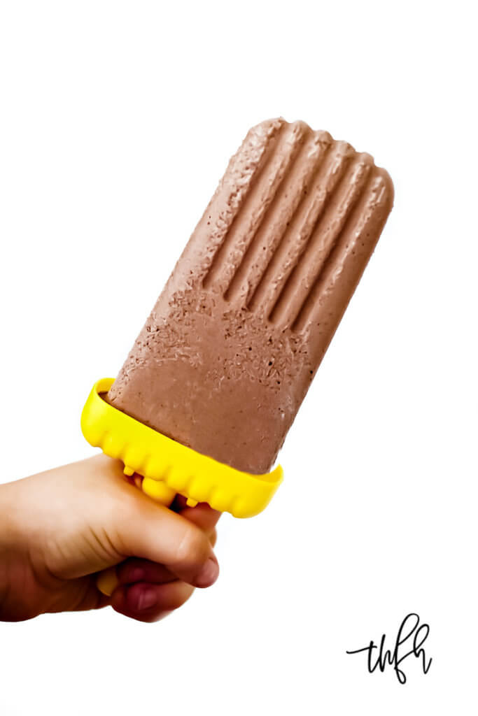 A small hand holding a chocolate fudgesicle on a solid white background