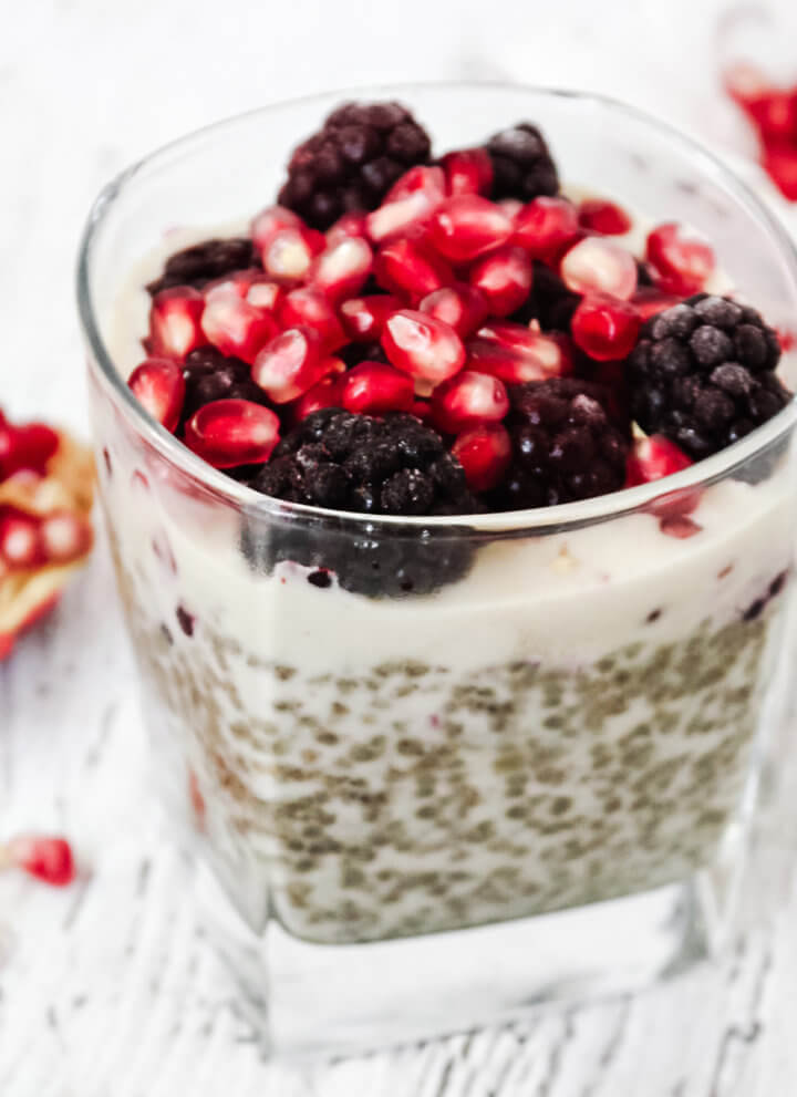 Square image of a square dessert glass filled with chia pudding topped with blackberries and pomegranates on a weathered wooden surface