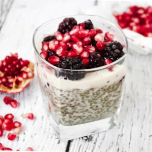 Horizontal image of a square dessert glass filled with chia pudding topped with blackberries and pomegranates on a weathered wooden surface