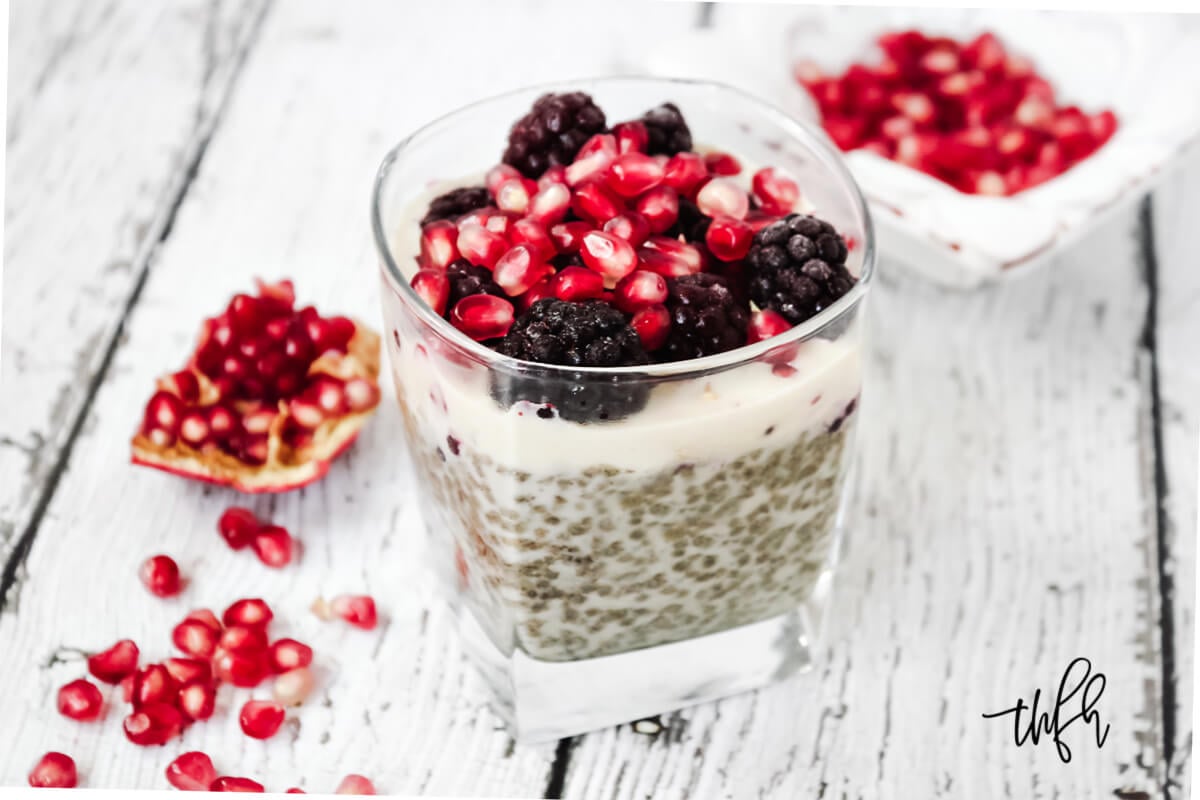 Horizontal image of a square dessert glass filled with chia pudding topped with blackberries and pomegranates on a weathered wooden surface
