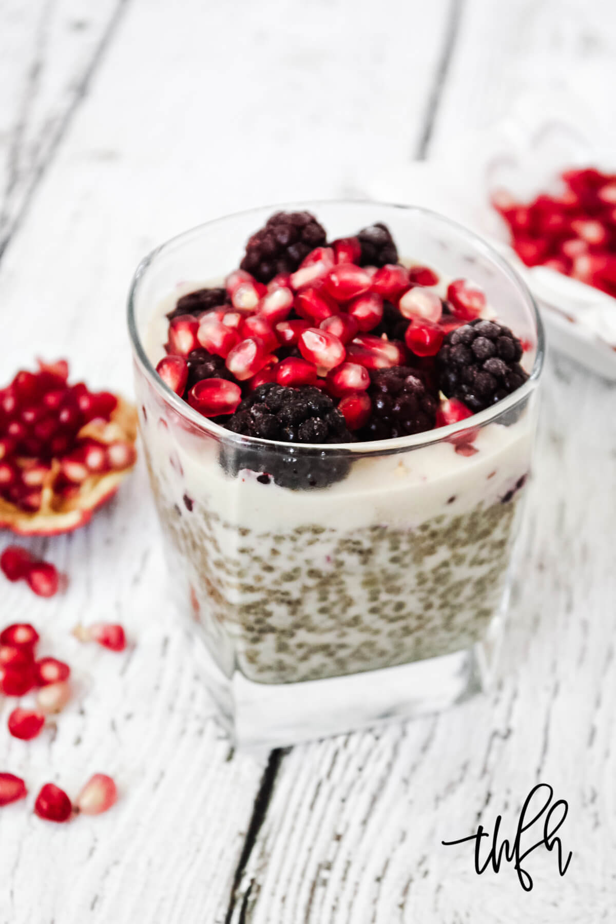 Vertical image of a square dessert glass filled with chia pudding topped with blackberries and pomegranates on a weathered wooden surface