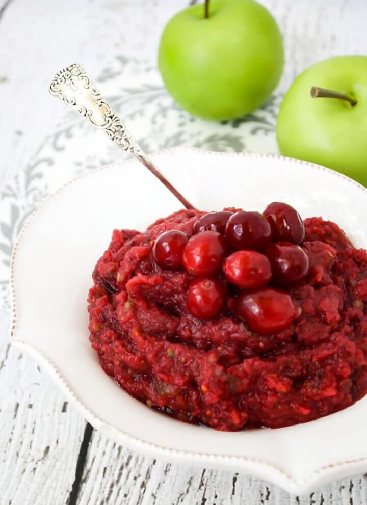 Vertical image of a decorative white bowl filled with no-cook cranberry sauce on a white weathered surface with two small green apples in the background