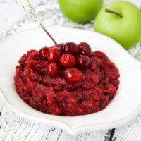 Horizontal image of a decorative white bowl filled with no-cook cranberry sauce on a white weathered surface with two small green apples in the background