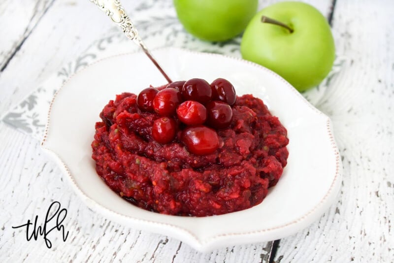 Horizontal image of a decorative white bowl filled with no-cook cranberry sauce on a white weathered surface with two small green apples in the background
