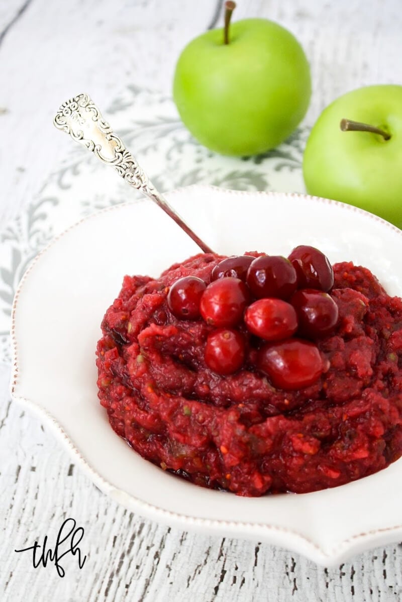 Vertical image of a decorative white bowl filled with no-cook cranberry sauce on a white weathered surface with two small green apples in the background
