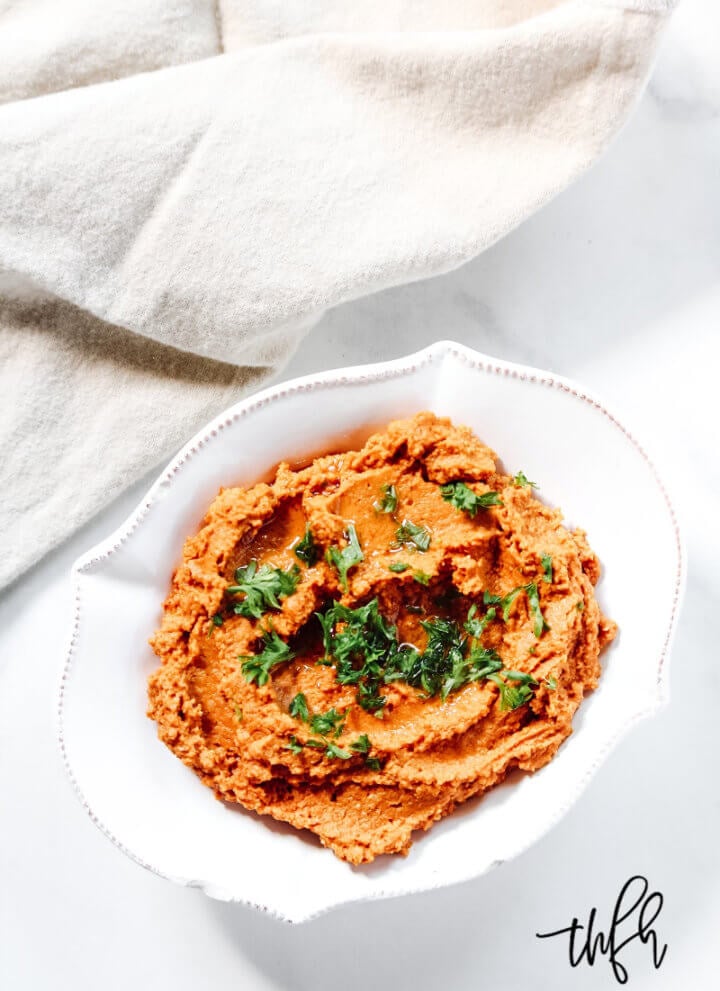 An overhead image of a decorative white bowl filled with Gluten-Free Vegan Smoky Chipotle Pumpkin Hummus with a cream napkin to the side on a solid white background