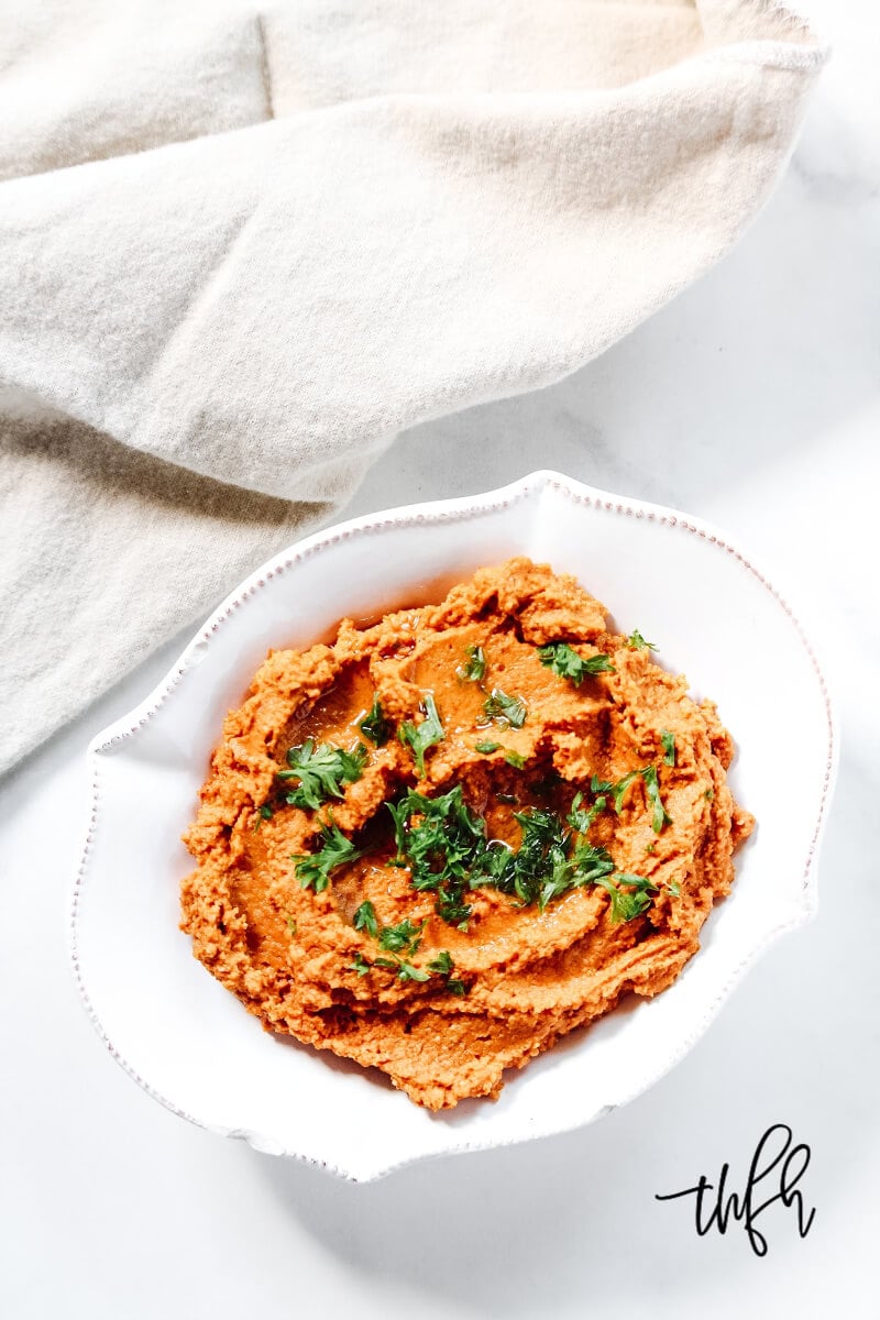 An overhead image of a decorative white bowl filled with Gluten-Free Vegan Smoky Chipotle Pumpkin Hummus with a cream napkin to the side on a solid white background