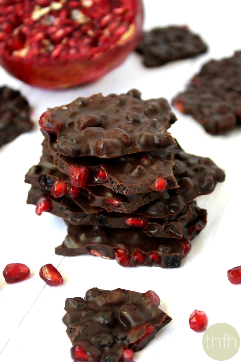 Stacked pieces of Clean Eating Gluten-Free Vegan No-Cook Pomegranate and Dried Blueberry Chocolate Bark on a white surface with a pomegranate fruit in the background