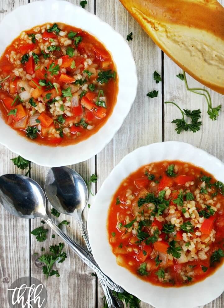 Vertical image of Vegan Stuffed Pepper Soup in two white bowls next to two spoons on a faded wooden surface with a loaf of bread in the background