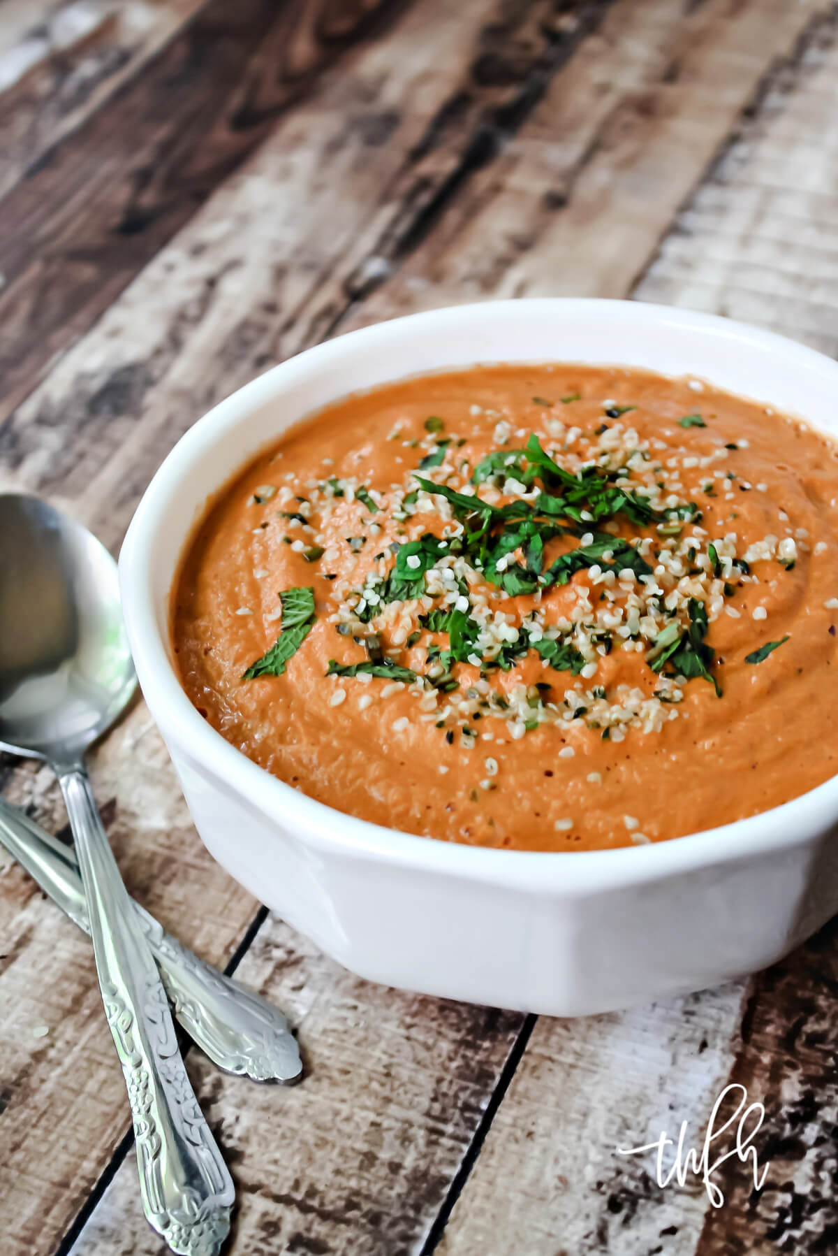 Vertical image of a white bowl filled with creamy tomato basil soup on top of a weathered wooden surface
