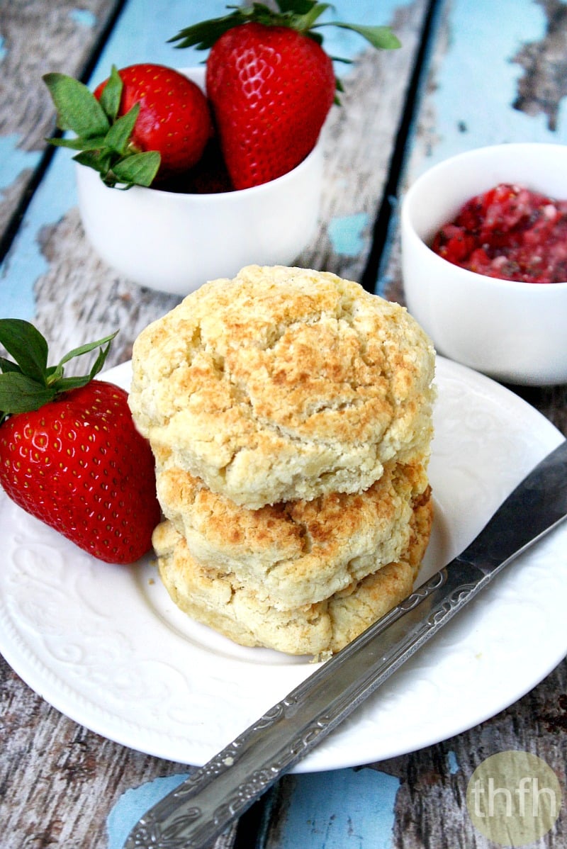 Vertical image of a stack of three Gluten-Free Vegan Biscuits on a white saucer surrounded by strawberries on a weathered wooden surface