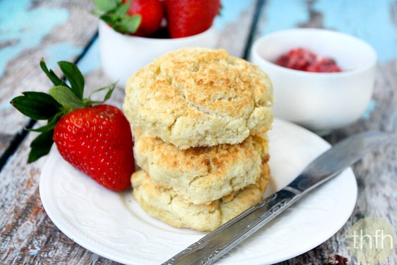 A stack of three Gluten-Free Vegan Biscuits on a white saucer surrounded by strawberries on a weathered wooden surface