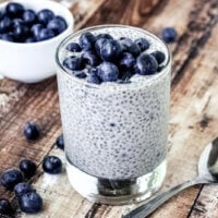 Vertical image of a glass filled with Vanilla Bean and Blueberry Chia Seed Pudding on a weathered wooden surface with a spoon and small bowl filled with blueberries to the side