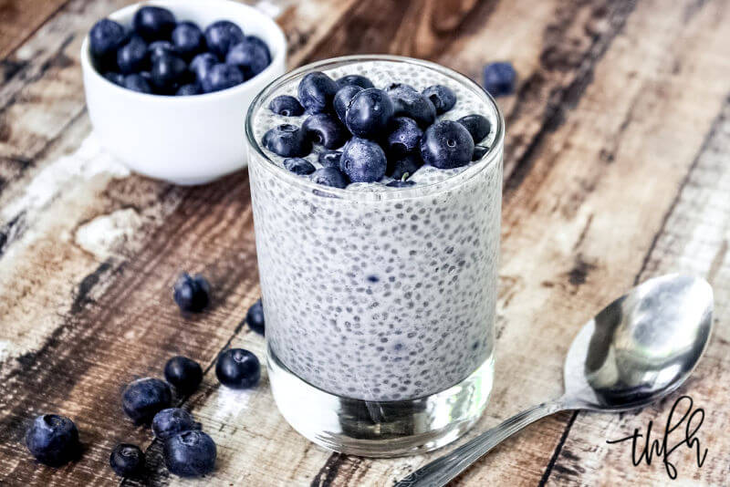 Vertical image of a glass filled with Vanilla Bean and Blueberry Chia Seed Pudding on a weathered wooden surface with a spoon and small bowl filled with blueberries to the side