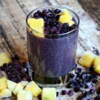 Wild Blueberry and Mango Protein Smoothie in a glass on a wooden surface with cubed mango and blueberries scattered