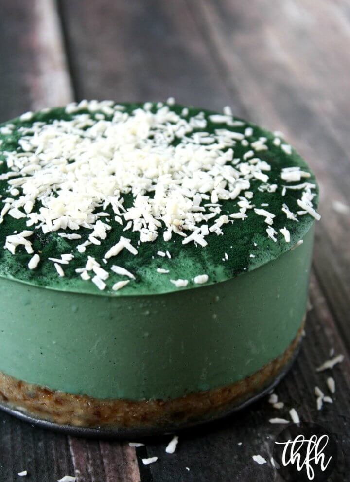 A vertical image of a whole uncut 6-inch Gluten-Free Vegan Raw No-Bake Spirulina Cheesecake sprinkled with shredded coconut on a weathered wooden surface