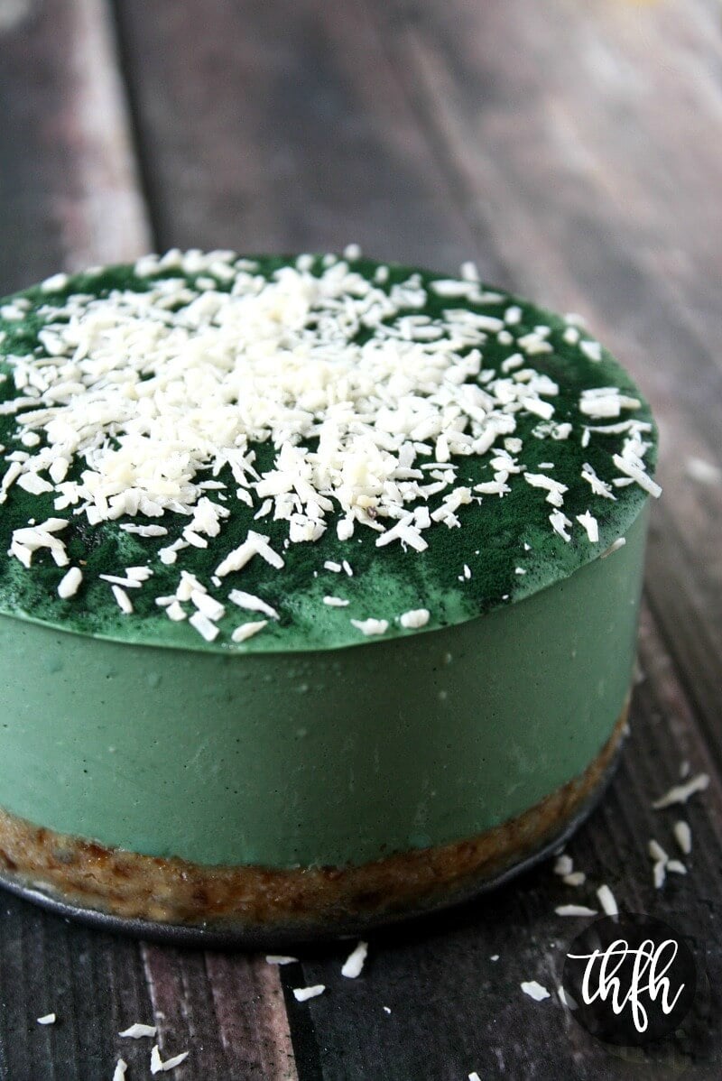 A vertical image of a whole 6-inch Gluten-Free Vegan Raw No-Bake Spirulina Cheesecake sprinkled with shredded coconut on a weathered wooden surface