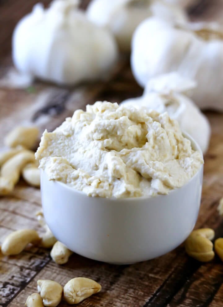 Vertical image of a small white dip bowl filled with Gluten-Free Vegan Garlic Cashew Spread with garlic bulbs scattered in the background