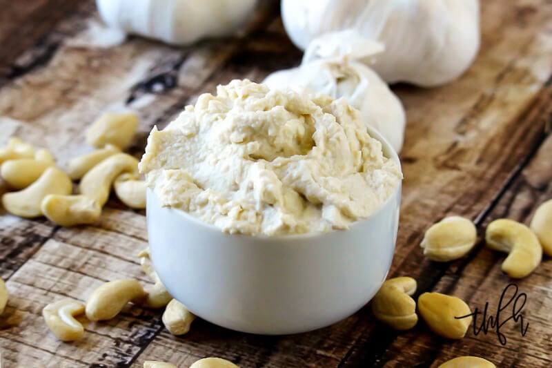 A small white bowl filled with Gluten-Free Vegan Garlic Cashew Spread on a weathered wooden surface with scattered cashews and garlic bulbs to the side