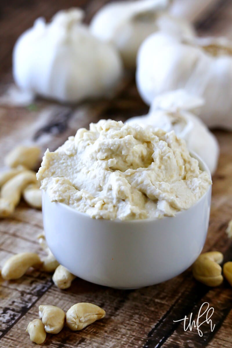 Vertical image of a small white dip bowl filled with Gluten-Free Vegan Garlic Cashew Spread on a weathered wooden surface with garlic bulbs in the background