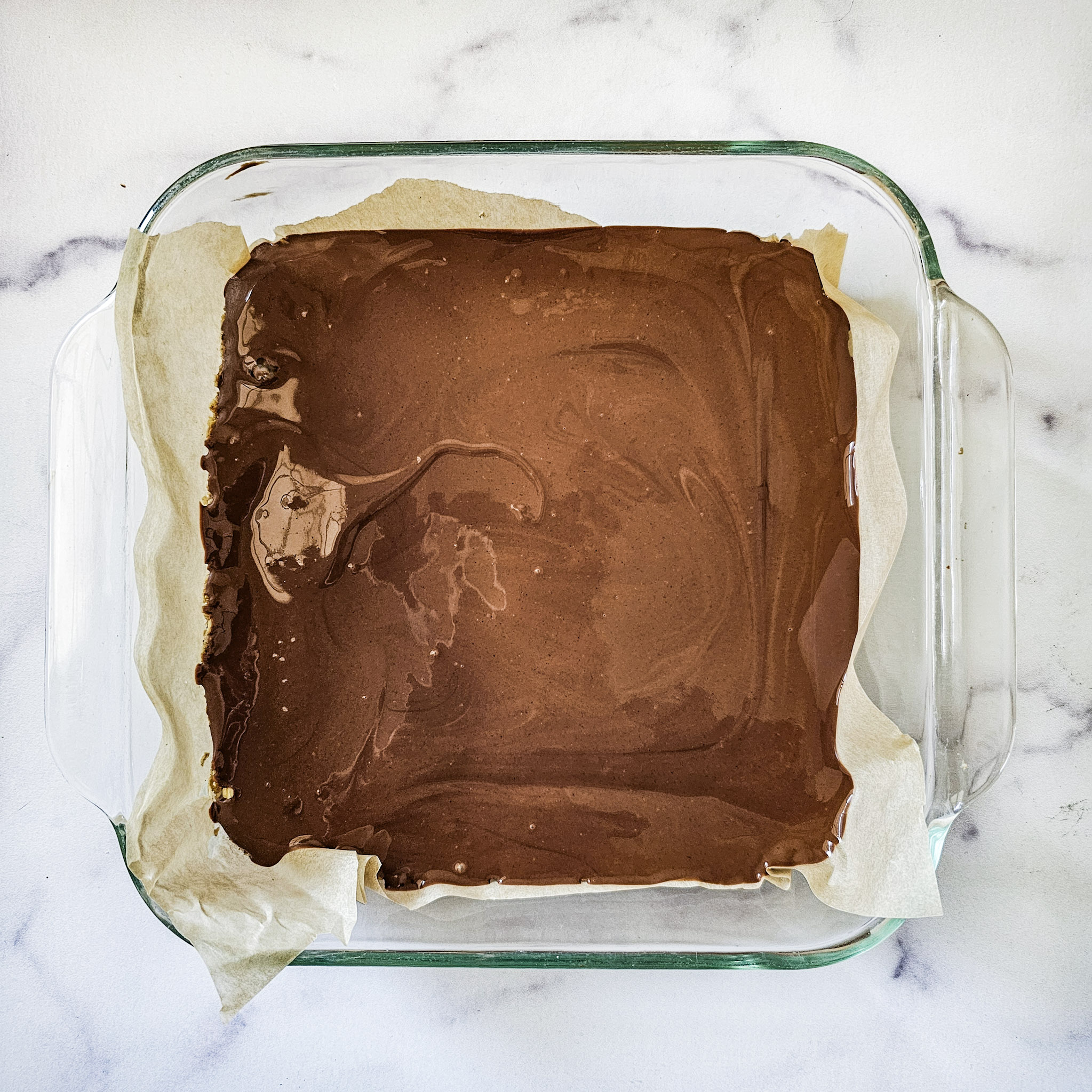 Overhead image of a chocolate peanut layer of gluten-free vegan no-bake chocolate peanut butter oat bars recipe in a glass baking dish lined with parchment paper on white marble background.