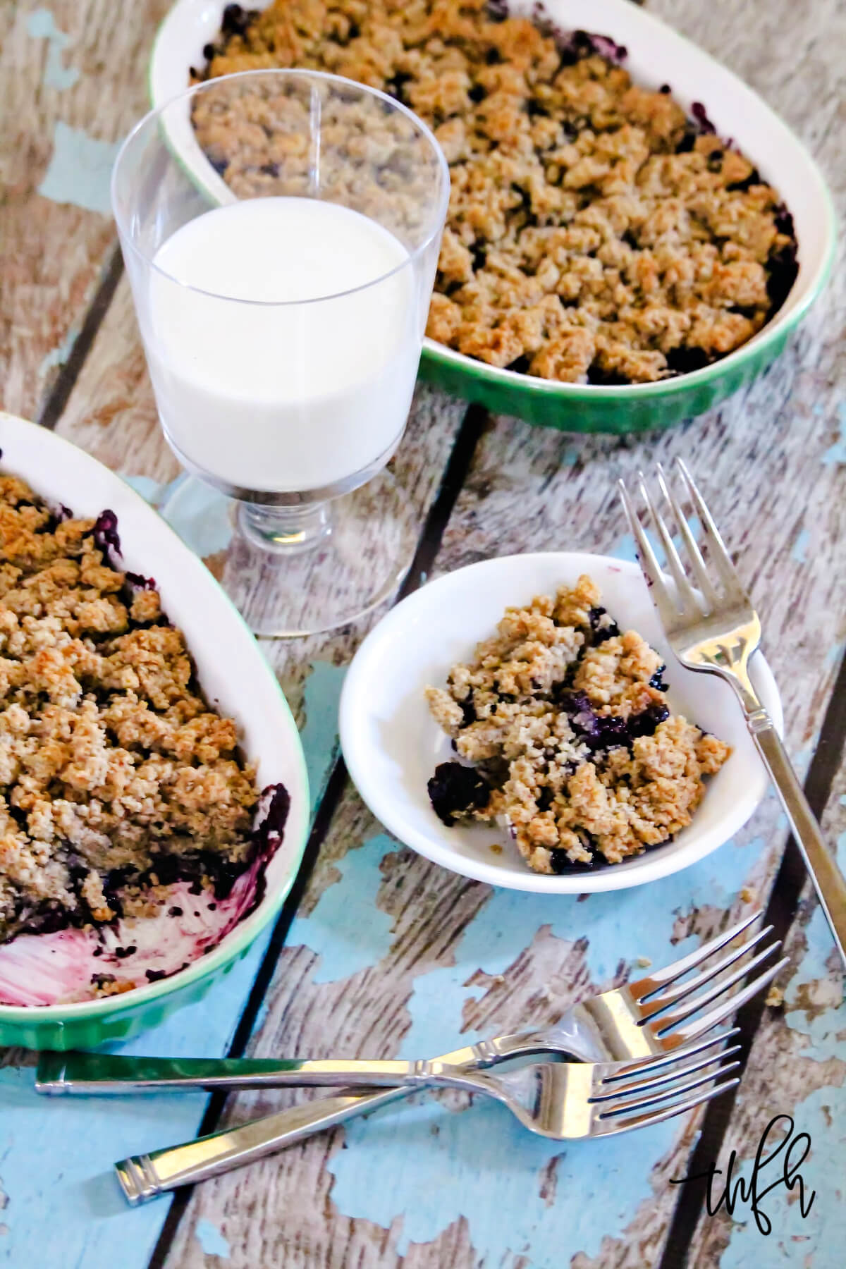 Overhead image of a small plate filled with blueberry cobbler with a glass of milk and several spoons to the side