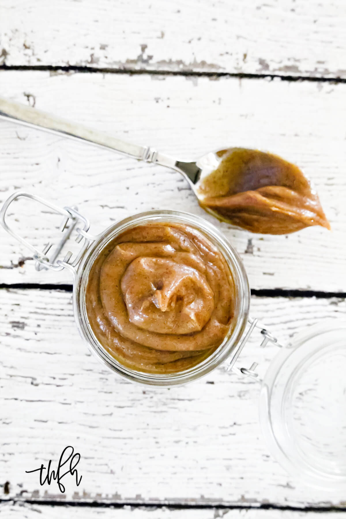 Vertical image of a small glass jar filled with homemade caramel on top of a weathered wooden surface