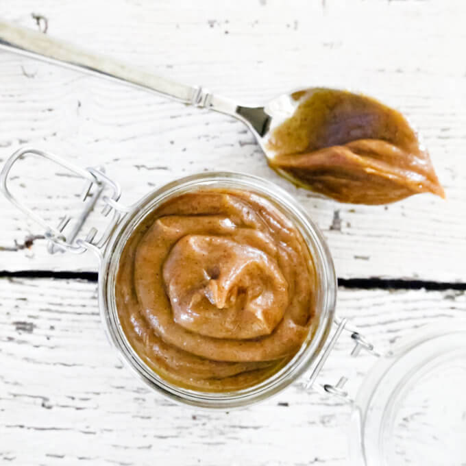 Square image of a small glass jar filled with homemade caramel on top of a weathered wooden surface