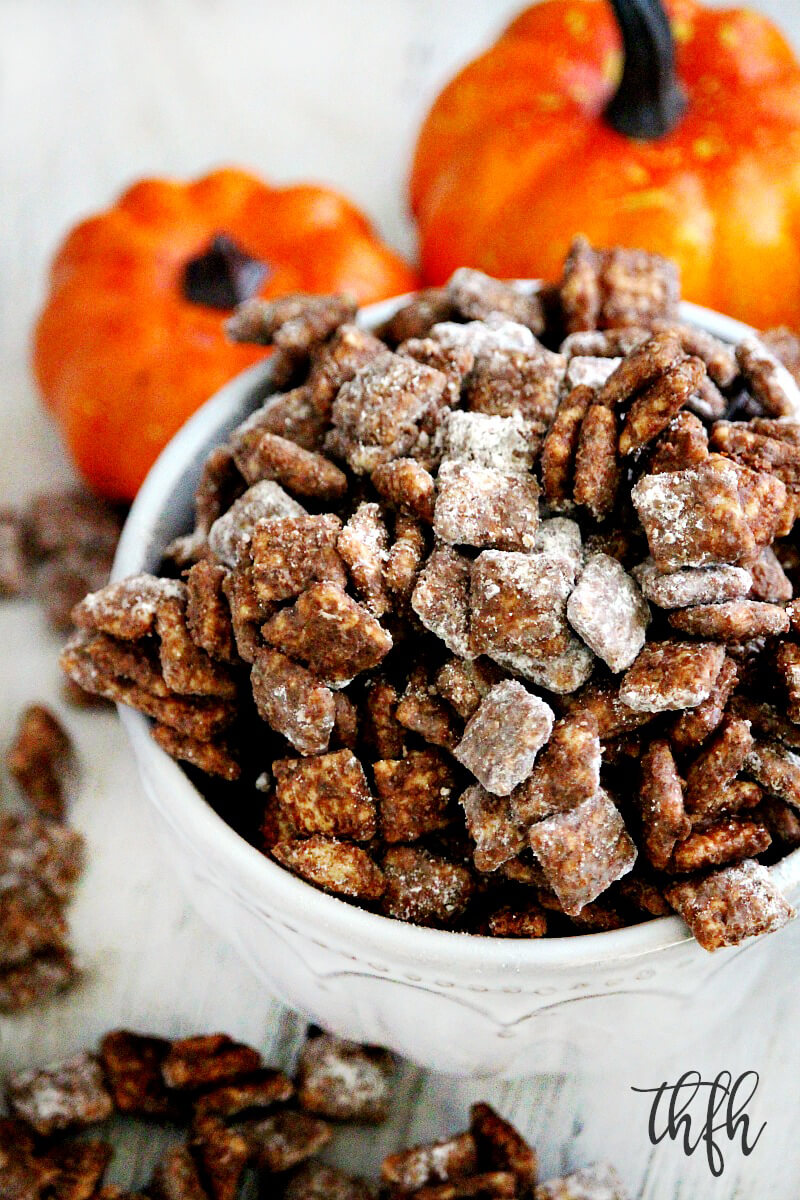 A close-up image of a white bowl filled and overflowing with Gluten-Free Vegan Pumpkin Spice "Puppy Chow" Muddy Buddies and two small pumpkins in the background