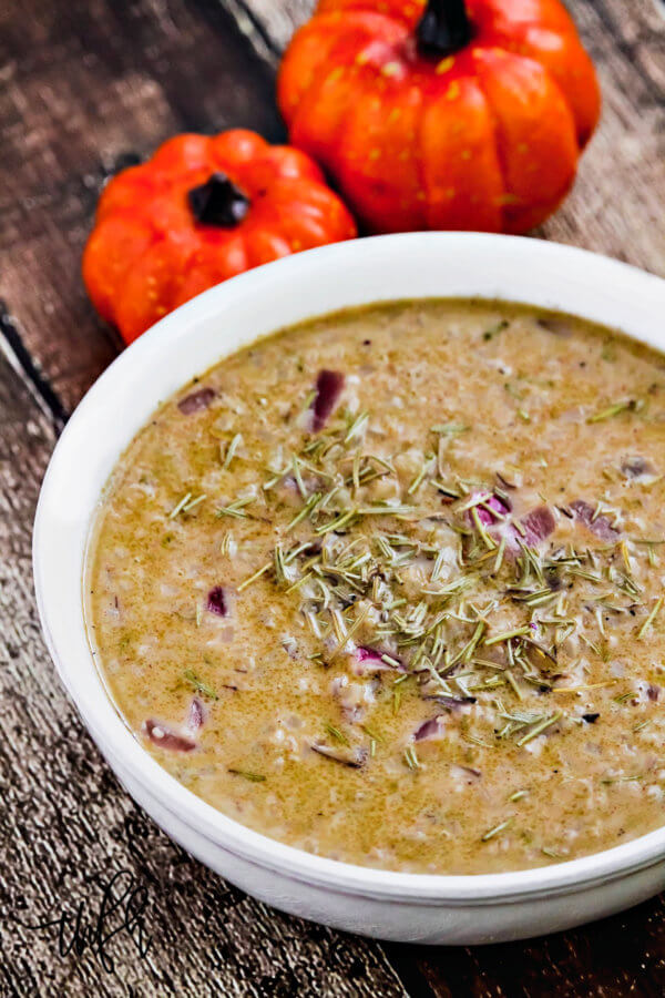 A vertical image of a white bowl filled with creamy Gluten-Free Vegan Pumpkin Spice Cauliflower Rice Soup on a weathered wooden surface with two small pumpkins in the background
