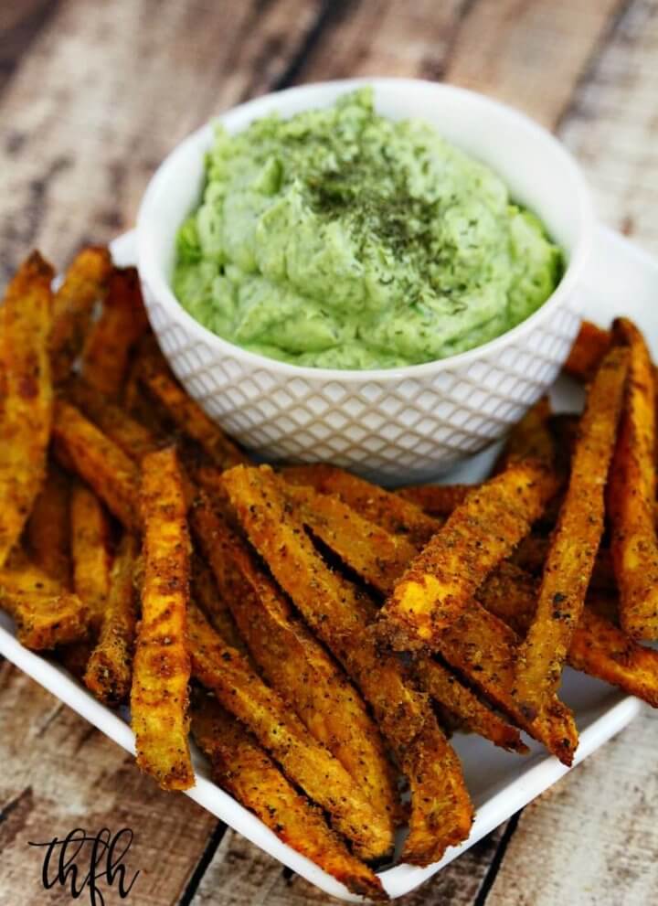Vertical image of a white plate filled with Lectin-Free Vegan Spicy Turmeric Oven-Baked Sweet Potato Fries next to a bowl of avocado dip on a weathered wooden surface