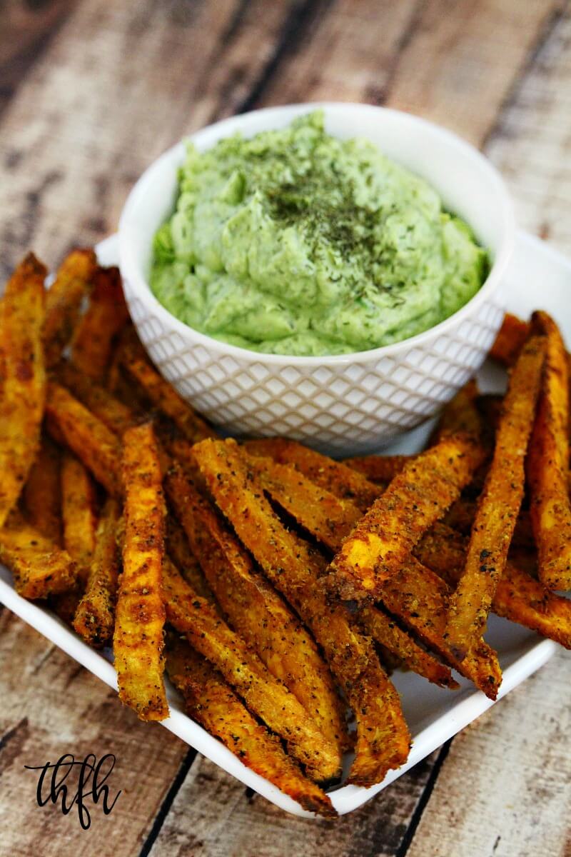 A close up image of a white plate filled with Lectin-Free Vegan Spicy Turmeric Oven-Baked Sweet Potato Fries next to a bowl of avocado dip on a weathered wooden surface